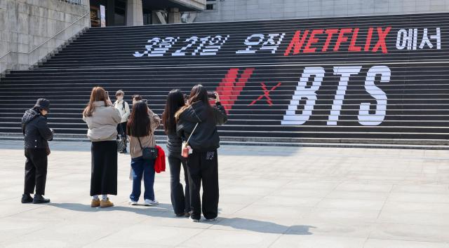 Tourists take a photo in front of a promotional banner for BTS’s comeback concert at the steps of Sejong Center for the Performing Arts in central Seoul on March 13. 2026. AJP Yoo Na-hyun 2026.03.13