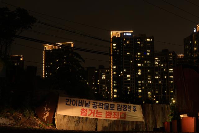 Lit apartment towers including one marked I PARK rise behind a corrugated metal fence bearing a protest banner reading in part Demolition is a crime near Guryong Village Seoul South Korea AJP Han Jun-gu