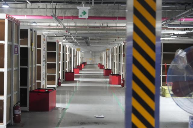 A long empty warehouse floor of Coupang with aisle lined alongside storage racks red bins and fire extinguishers marked with an exit sign 20260720 AJP Han Jun-gu