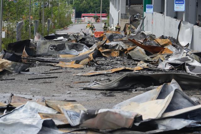 Firefighters in full gear walk past piles of burnt debris near the warehouses emergency assembly point signage with a fire truck visible on the road beyond AJP Han Jun-gu