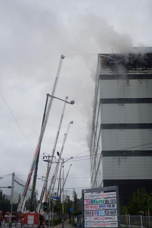 Fire trucks and aerial ladder units spray water onto the fire-damaged upper floors of the Coupang logistics center with dense smoke rising near the buildings rooftop sign AJP Han Jun-gu