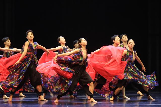An ensemble of dancers in black tops and flowing red skirts crouches low in unison arms curved overhead bathed in warm red stage lighting throughout the Seoul Metropolitan Dance Theatres Gut Whispering Steps AJP Han Jun-gu