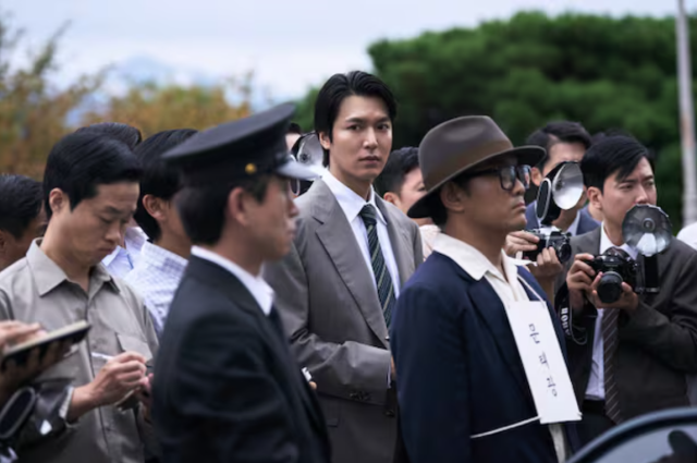 A scene from the South Korean film “The Assassin(s)” shows reporters and photographers gathered around a man during a tense public appearance. Courtesy of